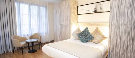 A hotel room with a large bed, a round table, two chairs, and a view of the outdoors through sheer curtains.