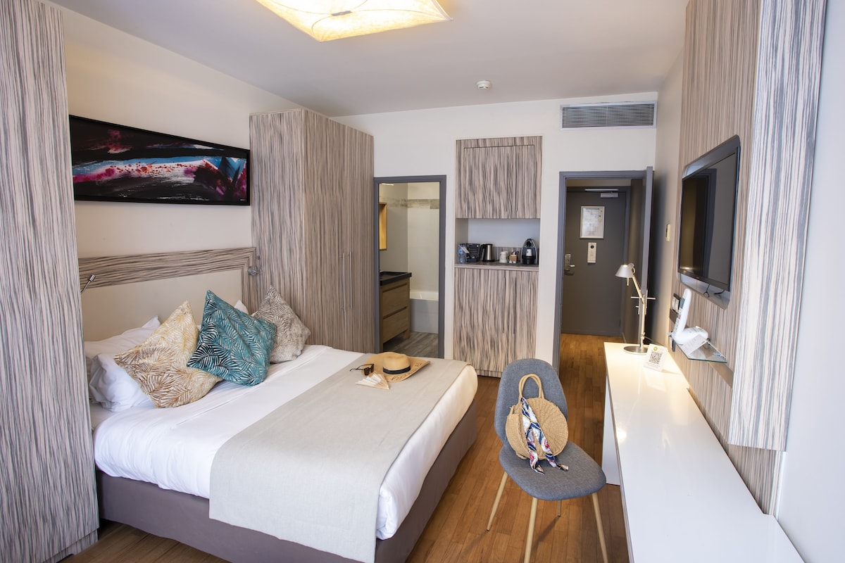 A modern hotel room with a bed, a desk, a chair, a TV, and a painting on the wall.