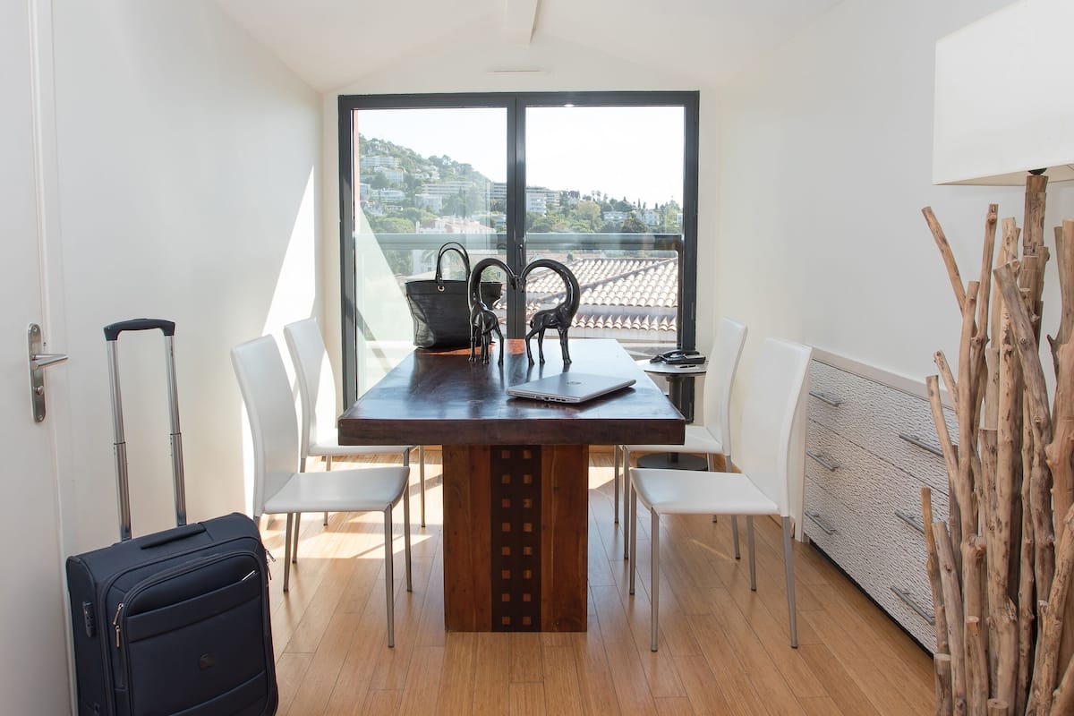 Une chambre moderne avec un plancher en bois, une table à manger avec des chaises, une grande fenêtre donnant sur un beau paysage, une valise et une structure décorative en bois.