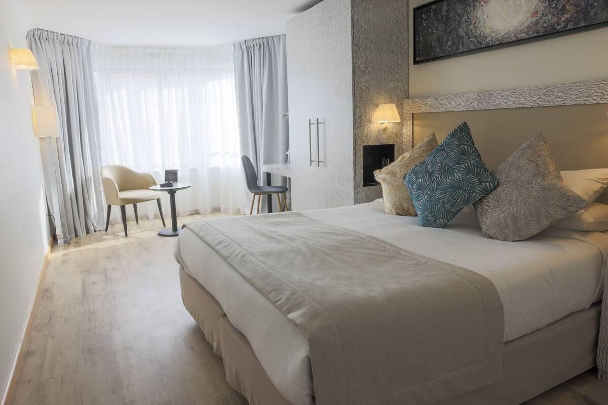 Une chambre d’hôtel avec un grand lit, un bureau et une chaise. Il y a un tableau au mur et une lampe sur le bureau.