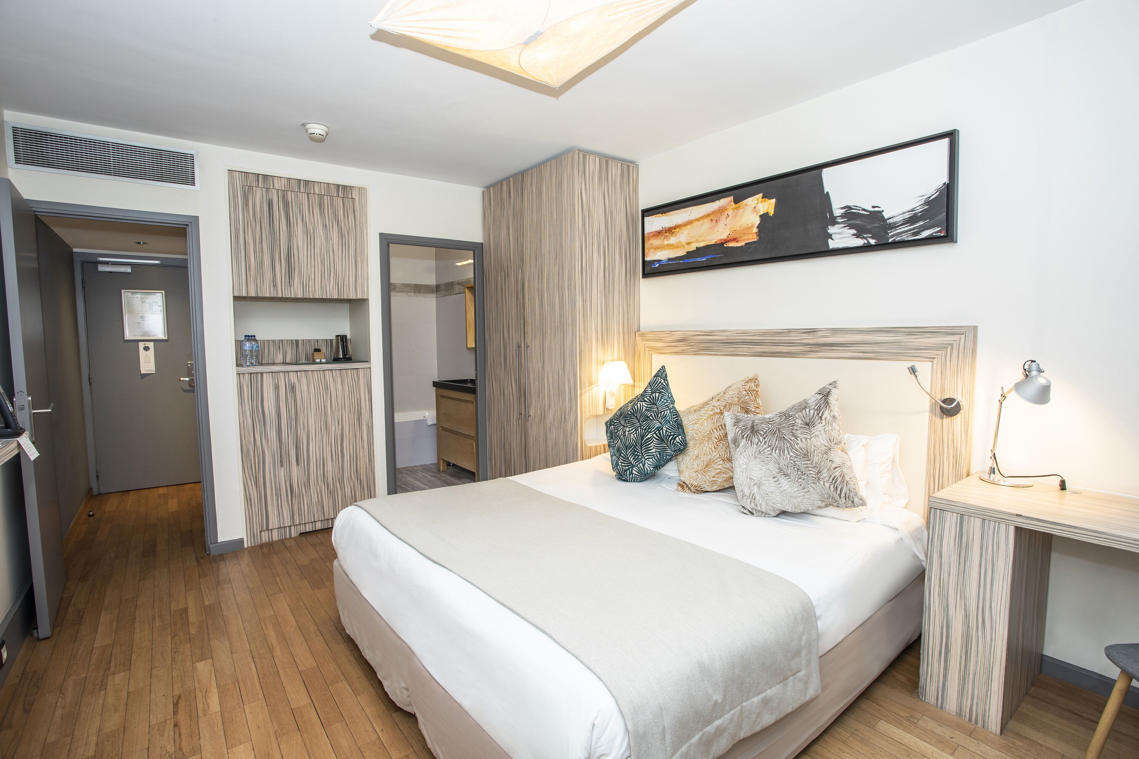 Una habitación de hotel moderna con una cama grande, cabecera de madera, mesita de noche con lámpara y un cuadro abstracto enmarcado en la pared.