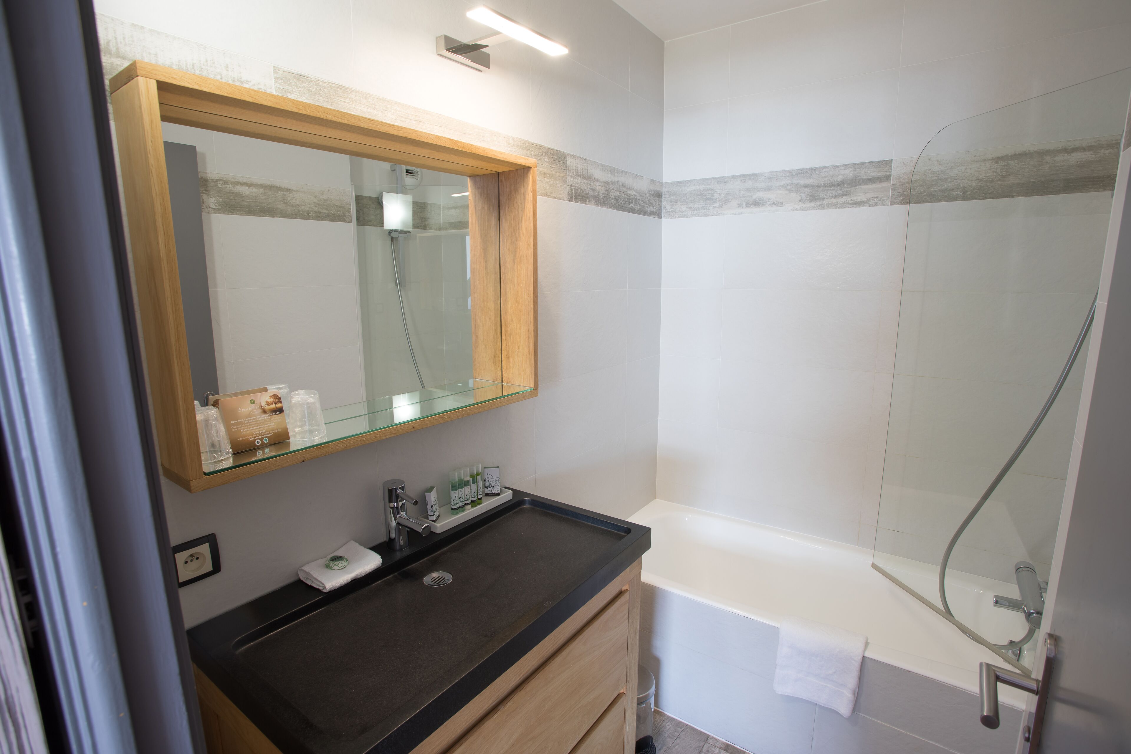Un baño moderno con cabina de ducha de vidrio, tocador de madera con encimera negra y bañera blanca.