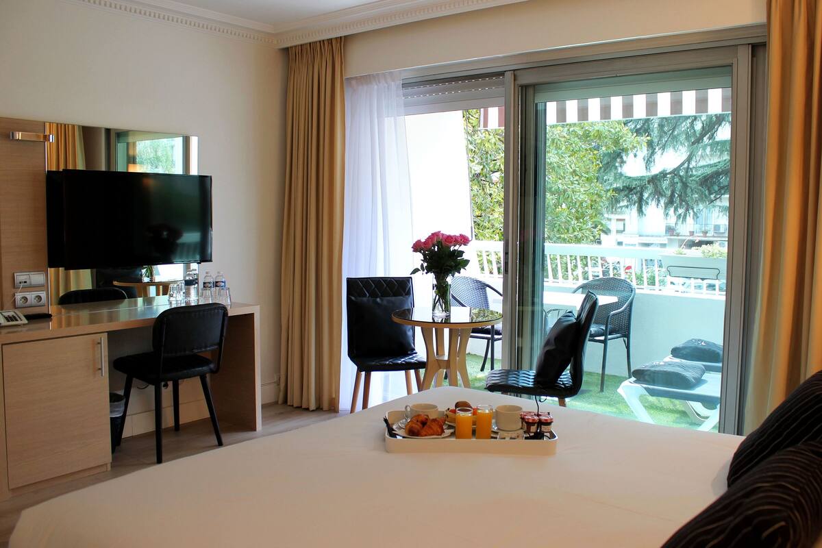 A hotel room with a bed, a desk with a TV, a dining area with chairs, and a balcony with a view.