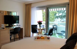 A hotel room with a bed, a desk with a TV, a dining area with chairs, and a balcony with a view.
