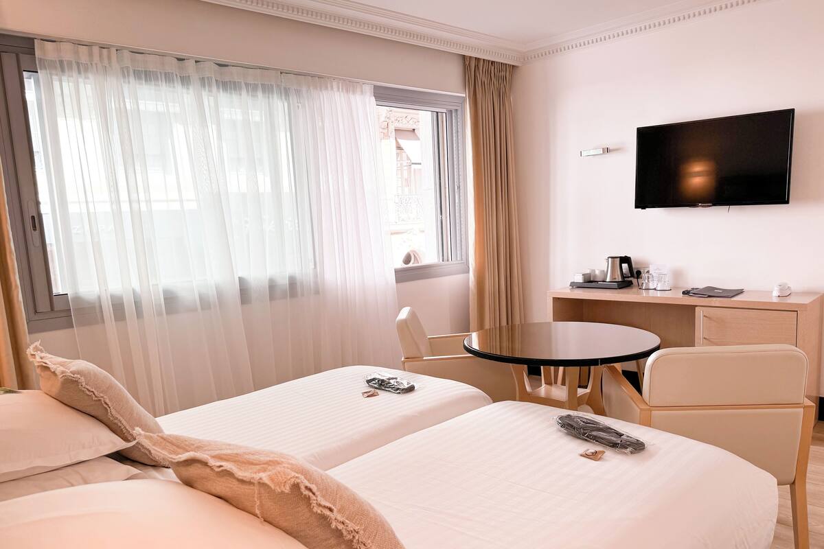 A hotel room with two beds, a desk, a chair, a television, and a window with sheer curtains.