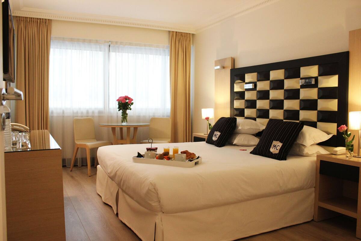 A hotel room with a large bed, a bedside table with a lamp, a small table with a vase of flowers, and a breakfast tray on the bed.