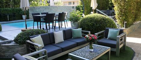 Un espace de détente aménagé au bord de la piscine, avec un canapé, des fauteuils et une table, le tout entouré de verdure et offrant une vue sur un bâtiment en arrière-plan.