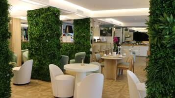 A modern indoor space with round tables and white chairs, surrounded by lush green walls and a wooden floor pattern.