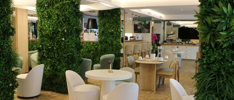 A modern indoor space with round tables and white chairs, surrounded by lush green walls and a wooden floor pattern.
