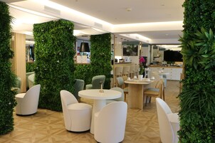 A modern indoor space with round tables and white chairs, surrounded by lush green walls and a wooden floor pattern.
