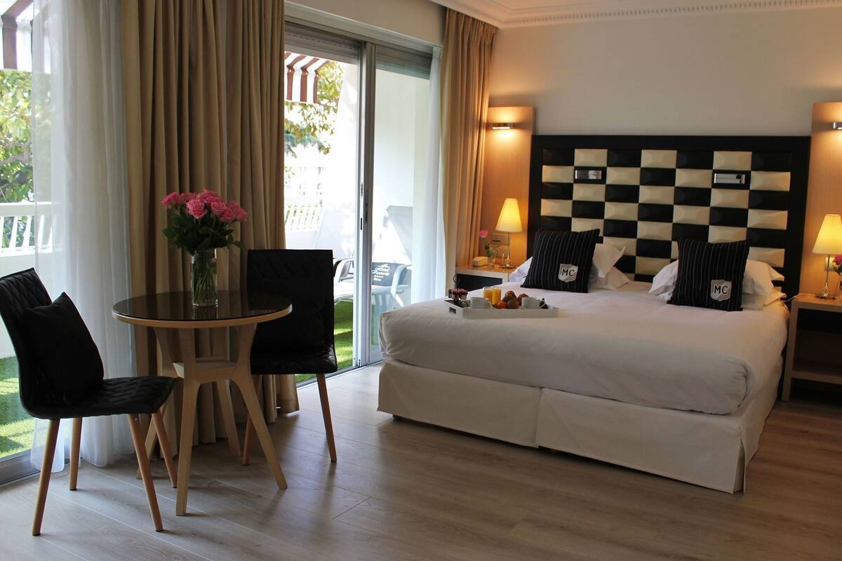 A hotel room with a bed, a table with chairs, a vase with flowers, and a view of the outdoors.