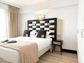 A hotel room with a large bed, a headboard with a checkered pattern, a bedside table with a lamp, and a view of the outside through the window.