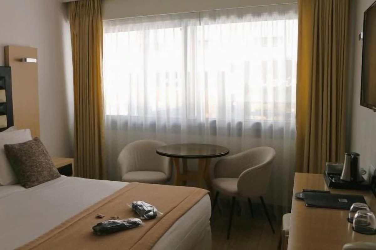 Hotellihuone, jossa on sänky, yöpöytä, tuoleja, pieni pöytä ja ikkuna, jossa on verhot.