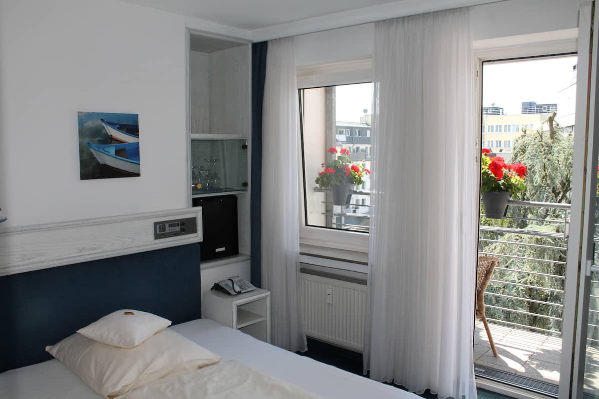 Una camera d'hotel con un letto, un comodino con un telefono, una finestra vista edifici e un balcone con piante in vaso.