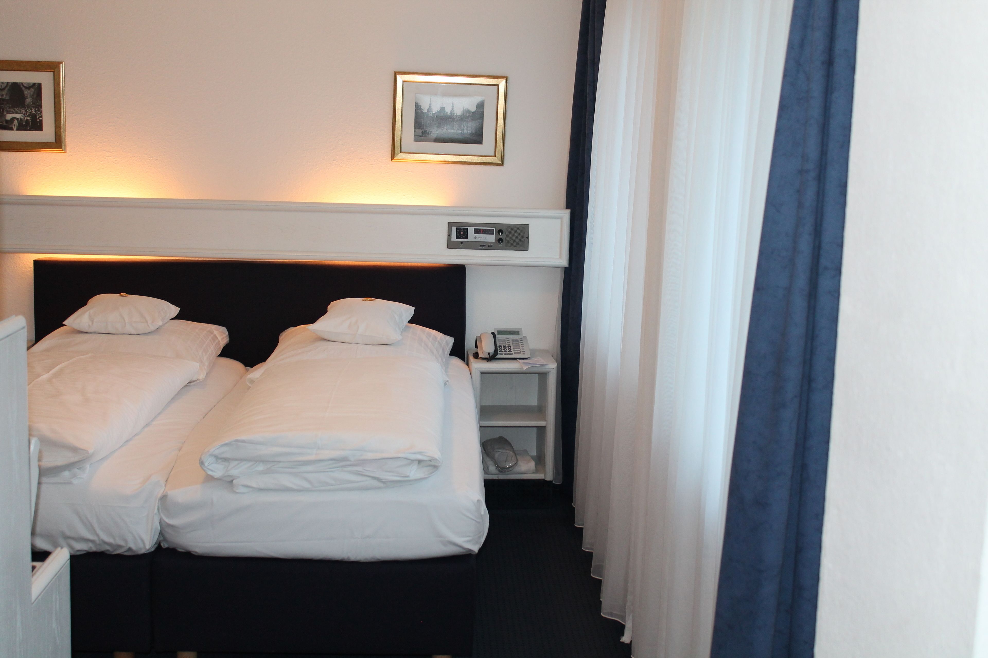 Habitación de hotel con dos camas, una mesita de noche con un teléfono, un estante pequeño y un cuadro en la pared.