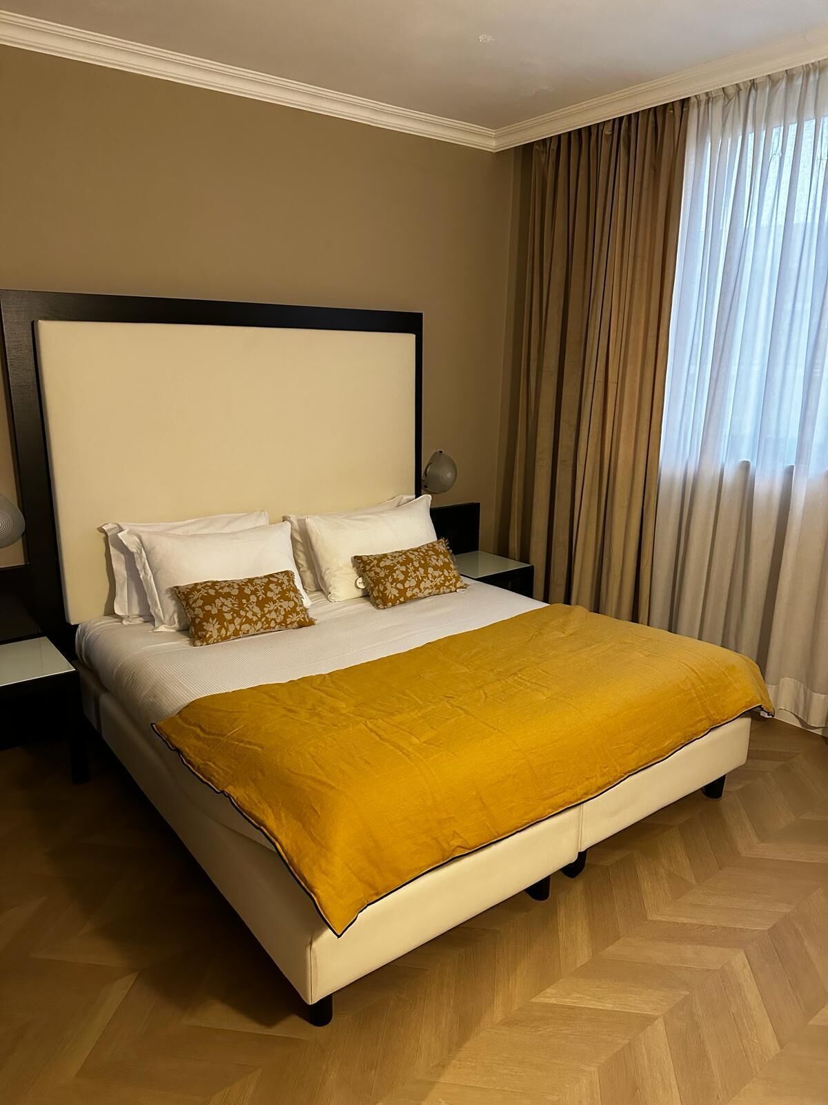 Una cama bien hecha con una manta color mostaza, un cabecero grande y dos mesitas de noche con lámparas.