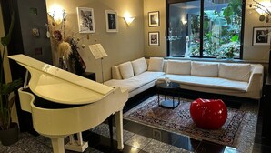 A modern living room with a white piano, a beige sofa, and a large window with a view of greenery. - Hotel Comtur (Binasco)