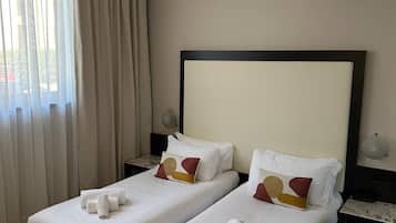 Habitación de hotel con dos camas, un ventanal con cortinas, una mesita de noche con lámpara y una alfombra en el suelo.