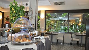 A buffet setup with covered food displays, a potted plant, and a view of a garden through a window. - Hotel Comtur (Binasco)
