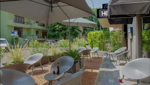 An outdoor seating area with modern chairs and tables, a covered area with umbrellas, and a building with a 'H' sign indicating a hotel. - Hotel Comtur (Binasco)