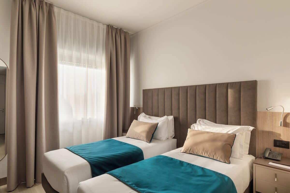 A hotel room with two beds, a large window with sheer curtains, a bedside table with a lamp, and a phone on the nightstand.