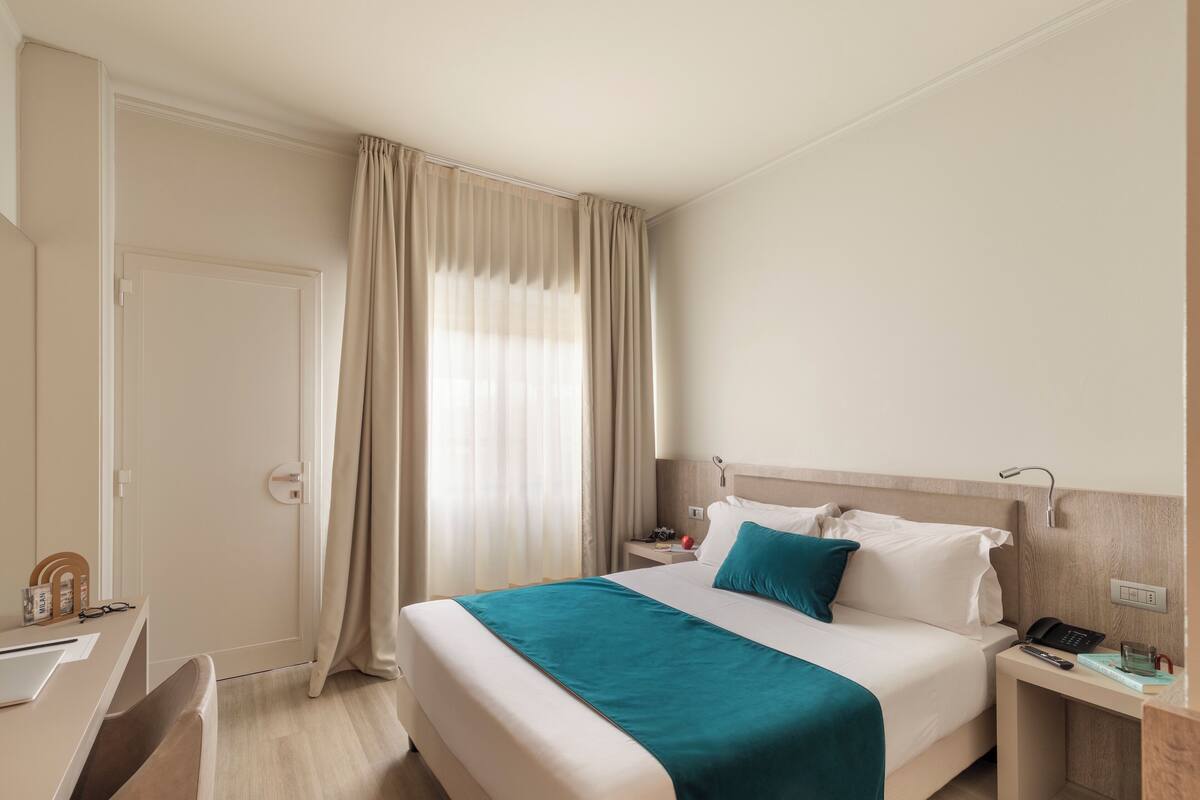 A hotel room with a bed, a desk, a chair, a nightstand, and a window with curtains.