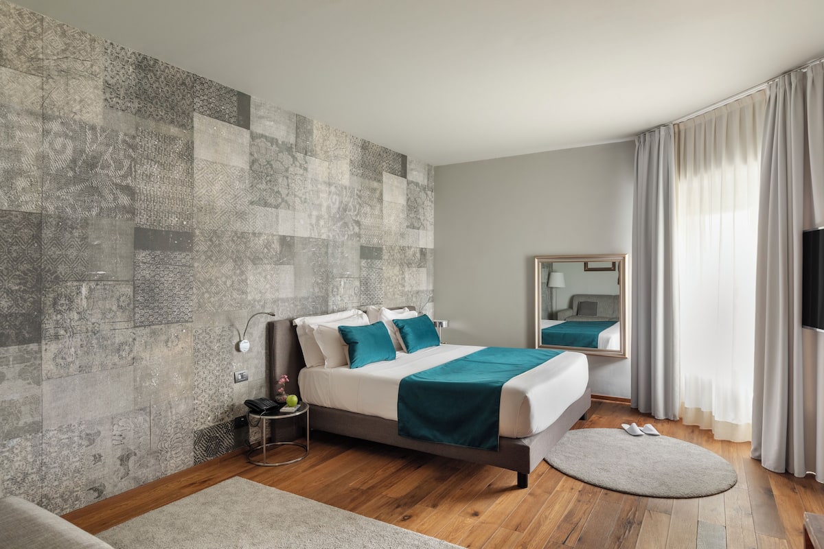 A modern hotel room with a large bed, a bedside table, a mirror, and a view of the outdoors.