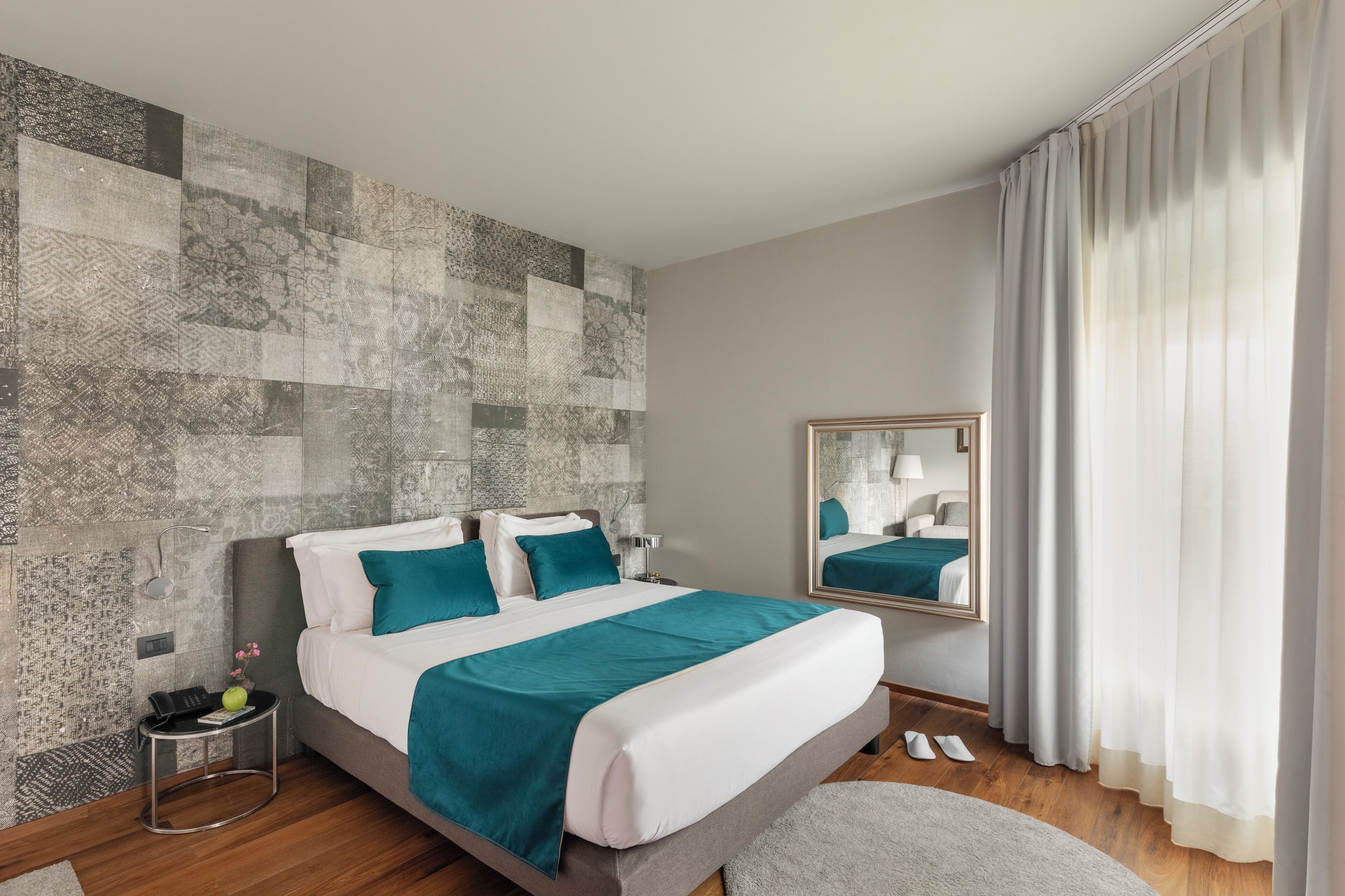 A modern hotel room with a large bed, a bedside table with a lamp, a mirror, and a view of the outdoors through sheer curtains.