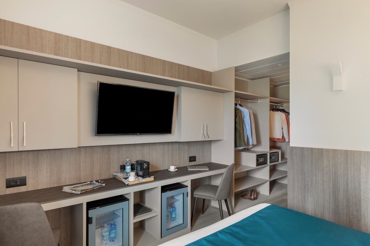 A modern hotel room with a flat-screen TV, a desk with a chair, a mini-fridge, a wardrobe, and a bed with a blue bedspread.