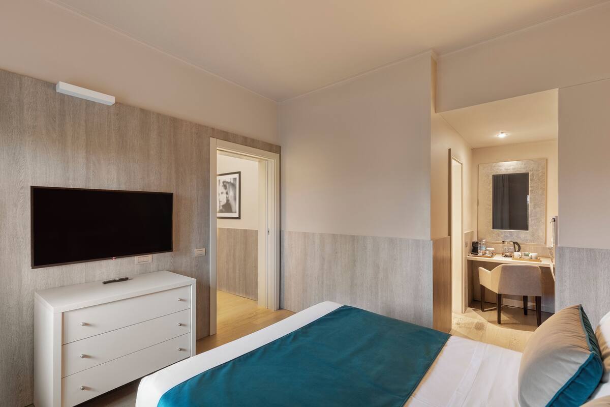 A modern hotel room with a flat-screen TV, a white dresser, a bed with teal bedding, a desk with a chair, and a bathroom with a mirror.