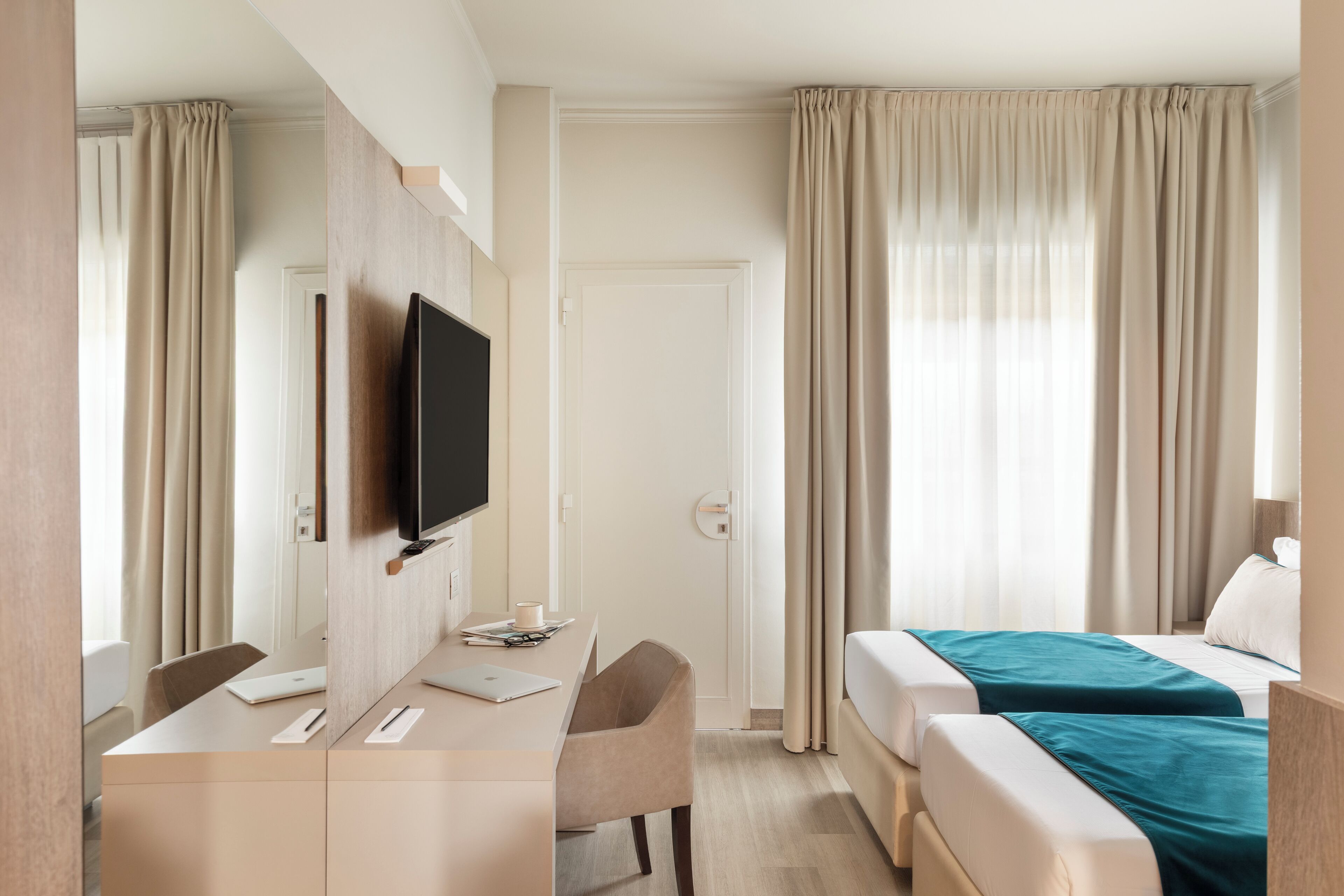 A hotel room with a bed, a desk with a computer, a TV, and a window with curtains.