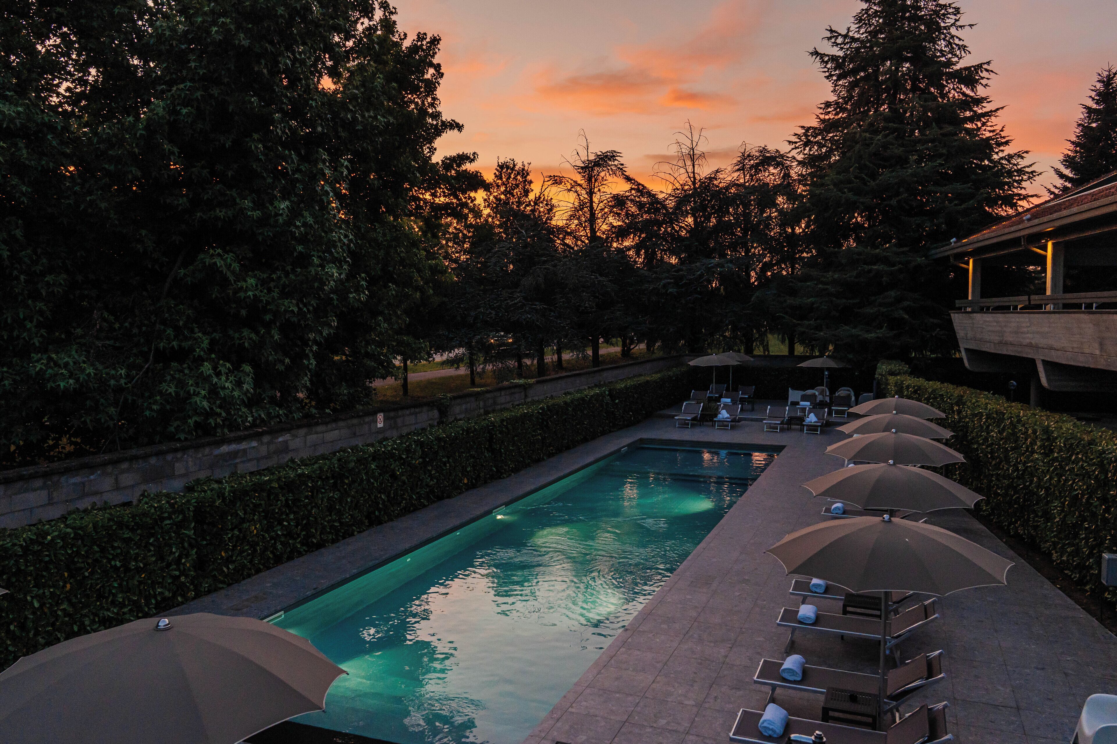 A swimming pool with lounge chairs and umbrellas, surrounded by trees and a building.