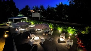 En utomhusplats med en terrass, vita loungestolar och bord, omgiven av grönska och upplyst av stämningsfull belysning.