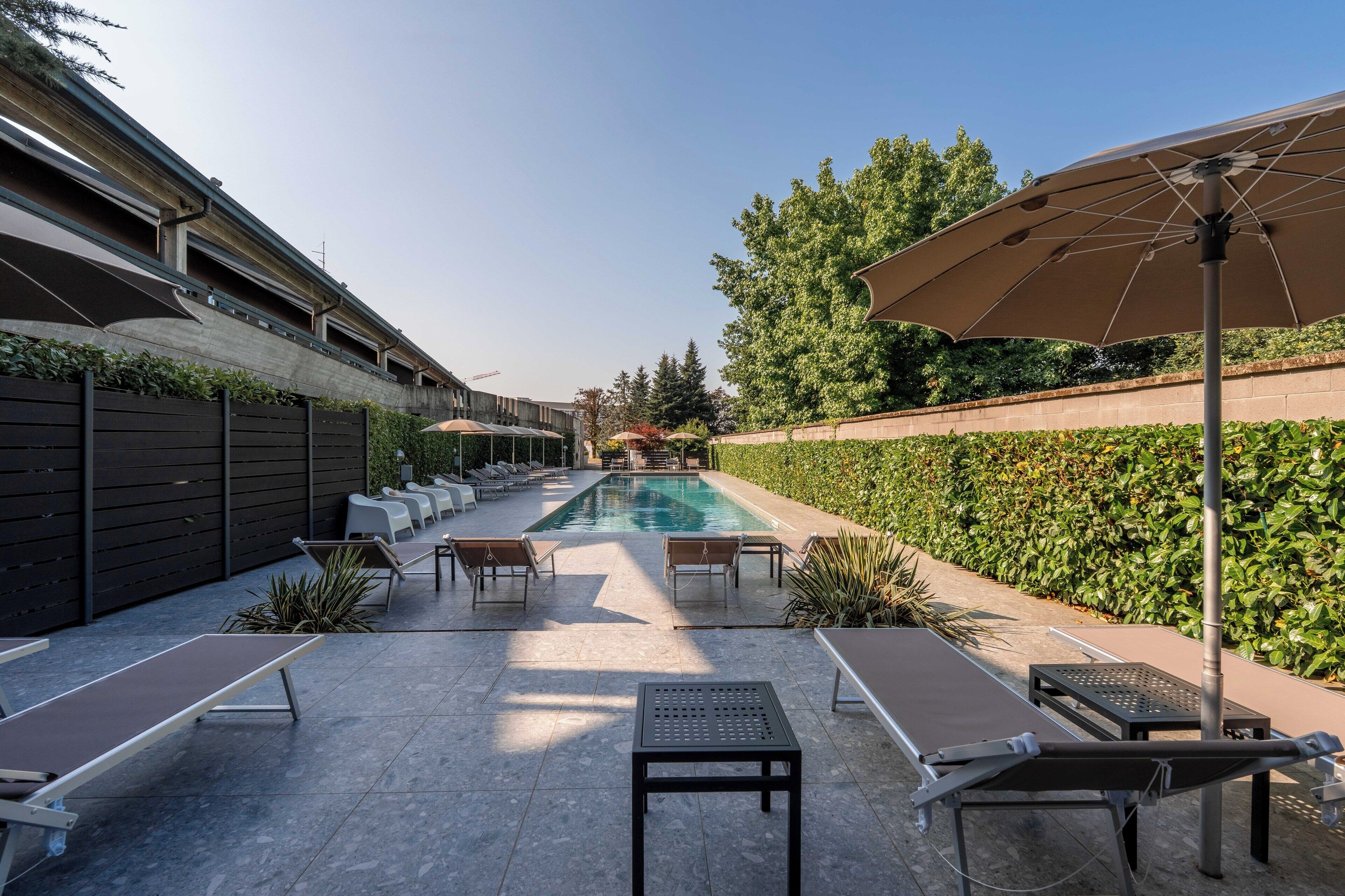 Zona de piscina con sillas de descanso y una mesa, rodeada por un seto y un edificio.