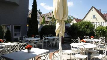 An outdoor patio with white metal tables and chairs, a yellow umbrella, and red flowers in vases.