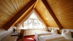A cozy attic bedroom with a sloped wooden ceiling, a large window, a desk, and a bed with pillows. - Airport Hotel Filderpost (Stuttgart)