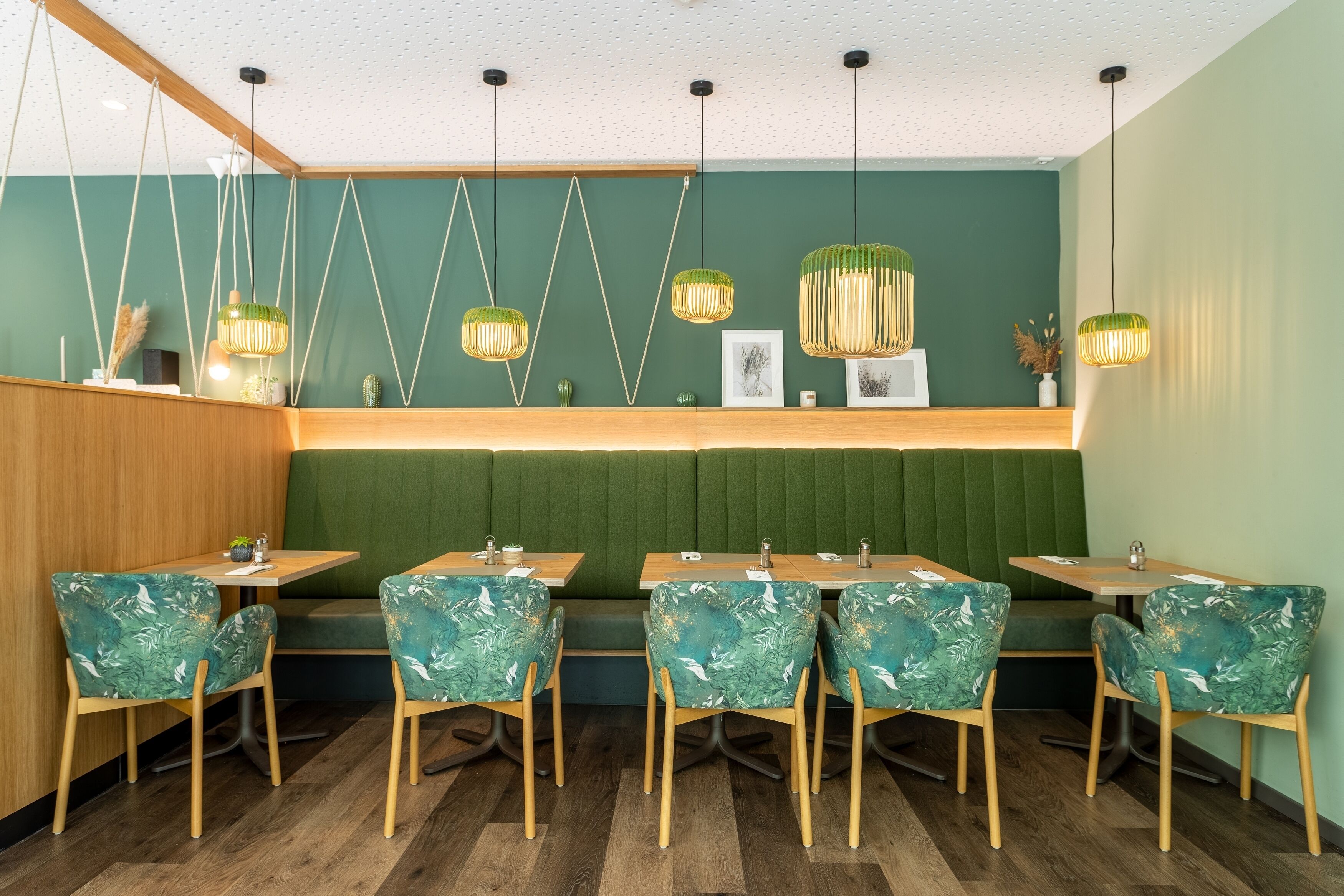 Interior de restaurante con mesas y sillas de madera, cabinas tapizadas de verde, lámparas colgantes y una pared verde con un panel de madera a la izquierda.