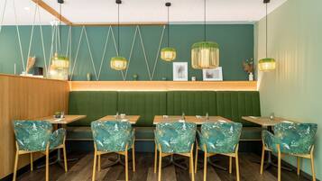 Interior de restaurante con mesas y sillas de madera, cabinas tapizadas de verde, lámparas colgantes y una pared verde con un panel de madera a la izquierda.