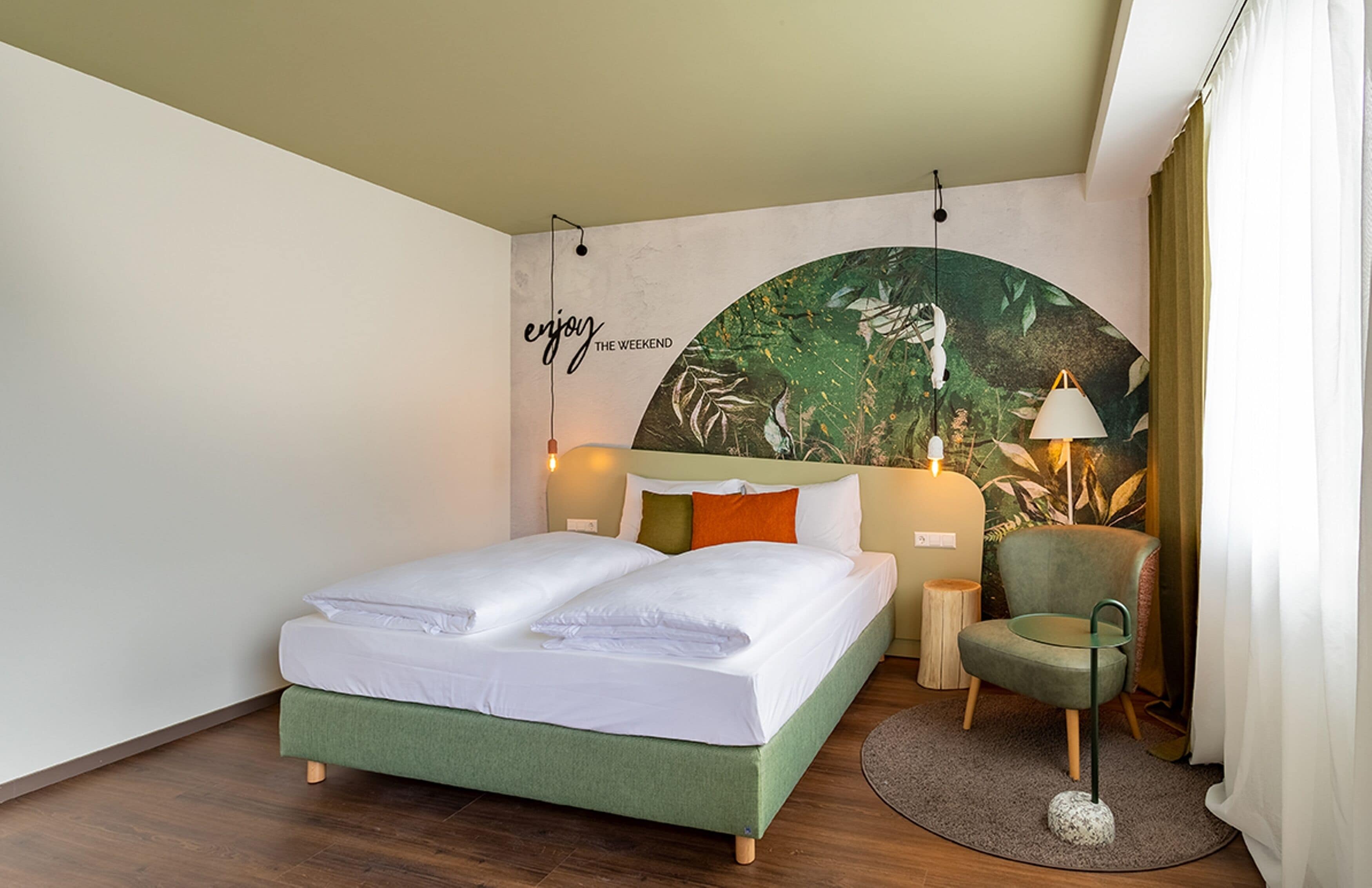 Habitación de hotel con una cama, una silla, una lámpara y un mural tropical en la pared.