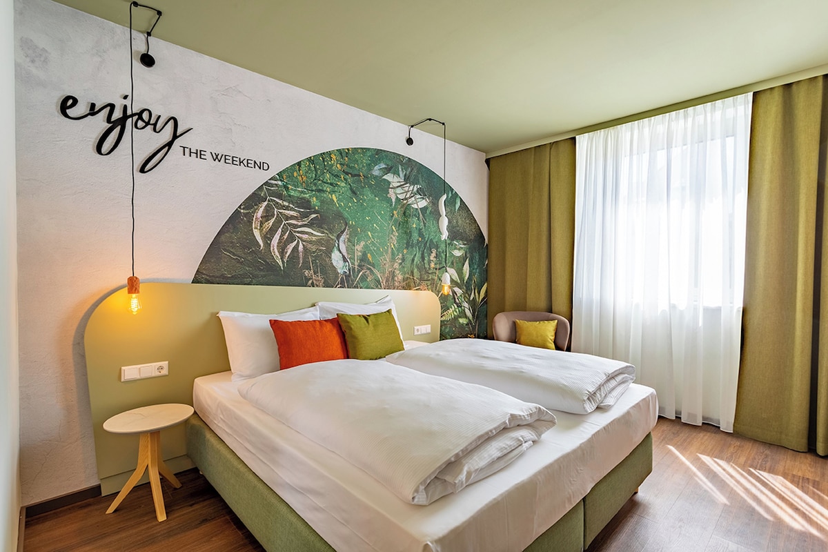 Una habitació d'hotel amb un llit, una taula petita, una cadira, una paret amb un mural de temàtica natural i una finestra gran amb cortines.