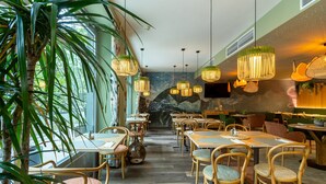 Um restaurante com mesas e cadeiras de madeira, luminárias pendentes e uma grande planta.