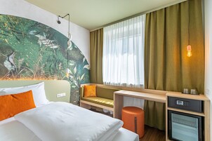 Quarto de hotel com cama, escrivaninha, cadeira, mesinha, painel decorativo na parede, janela com cortinas e luminária fixada na parede.