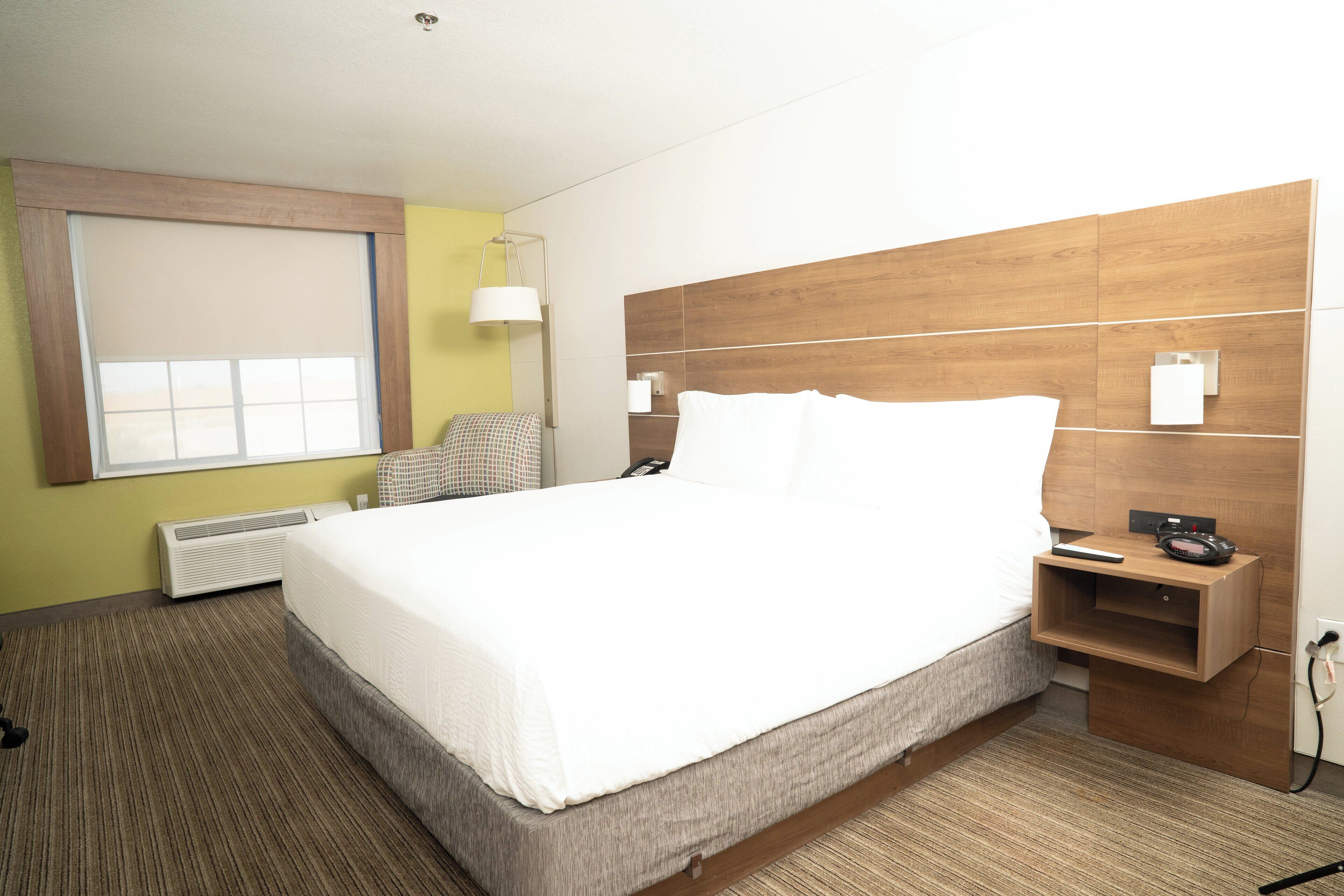 A hotel room with a large bed, a bedside table with a lamp, a chair, and a window with blinds.