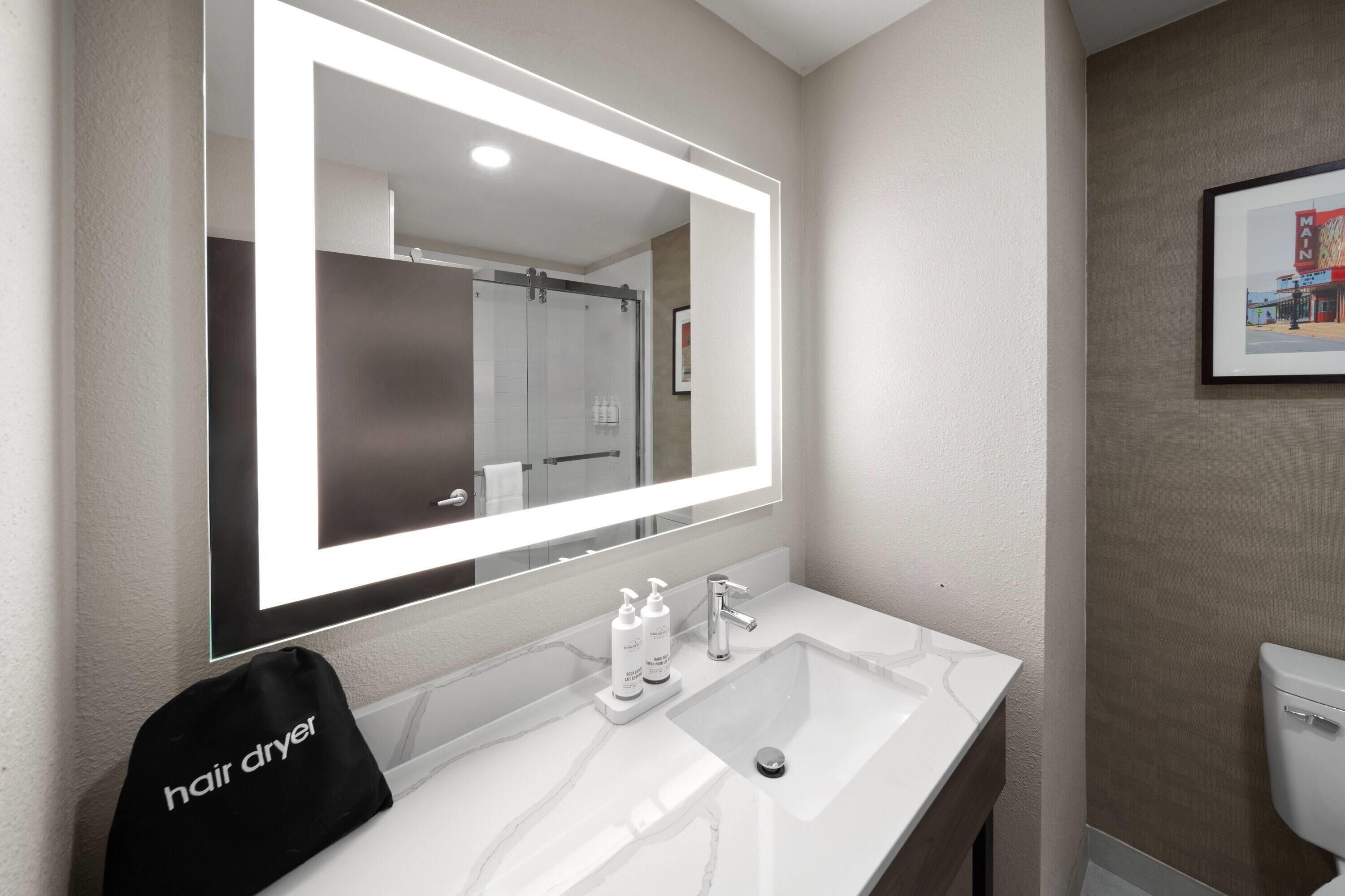 Une salle de bain avec un grand miroir, un lavabo blanc, un sèche-cheveux et un tableau encadré au mur.