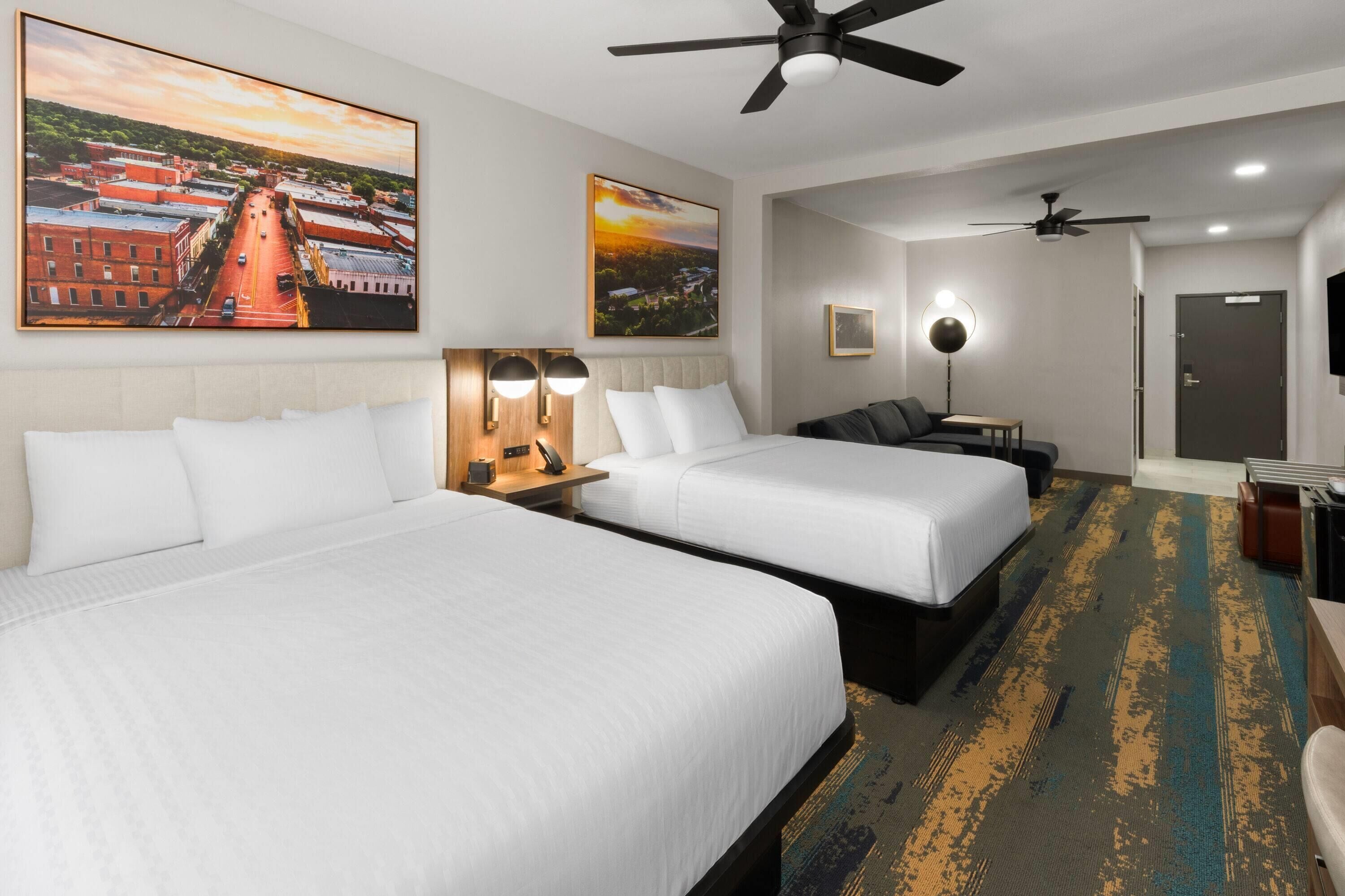 Quarto de hotel com duas camas, uma escrivaninha, uma cadeira, um ventilador de teto e uma grande pintura de uma paisagem urbana.