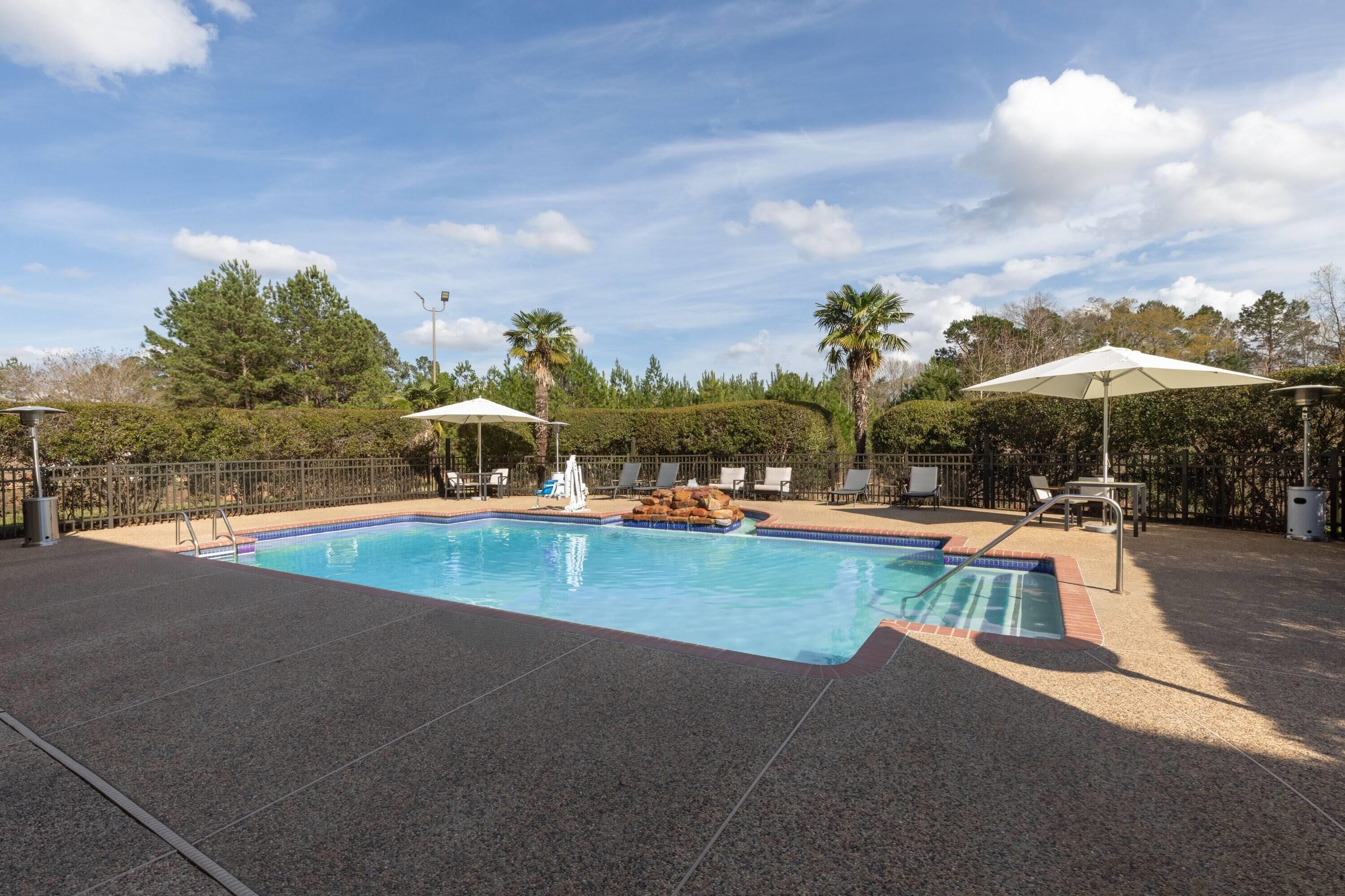 A swimming pool with a tiled deck, lounge chairs, umbrellas, and a fenced area with trees in the background.