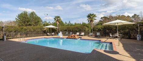 A swimming pool with a tiled deck, lounge chairs, umbrellas, and a fenced area with trees in the background.