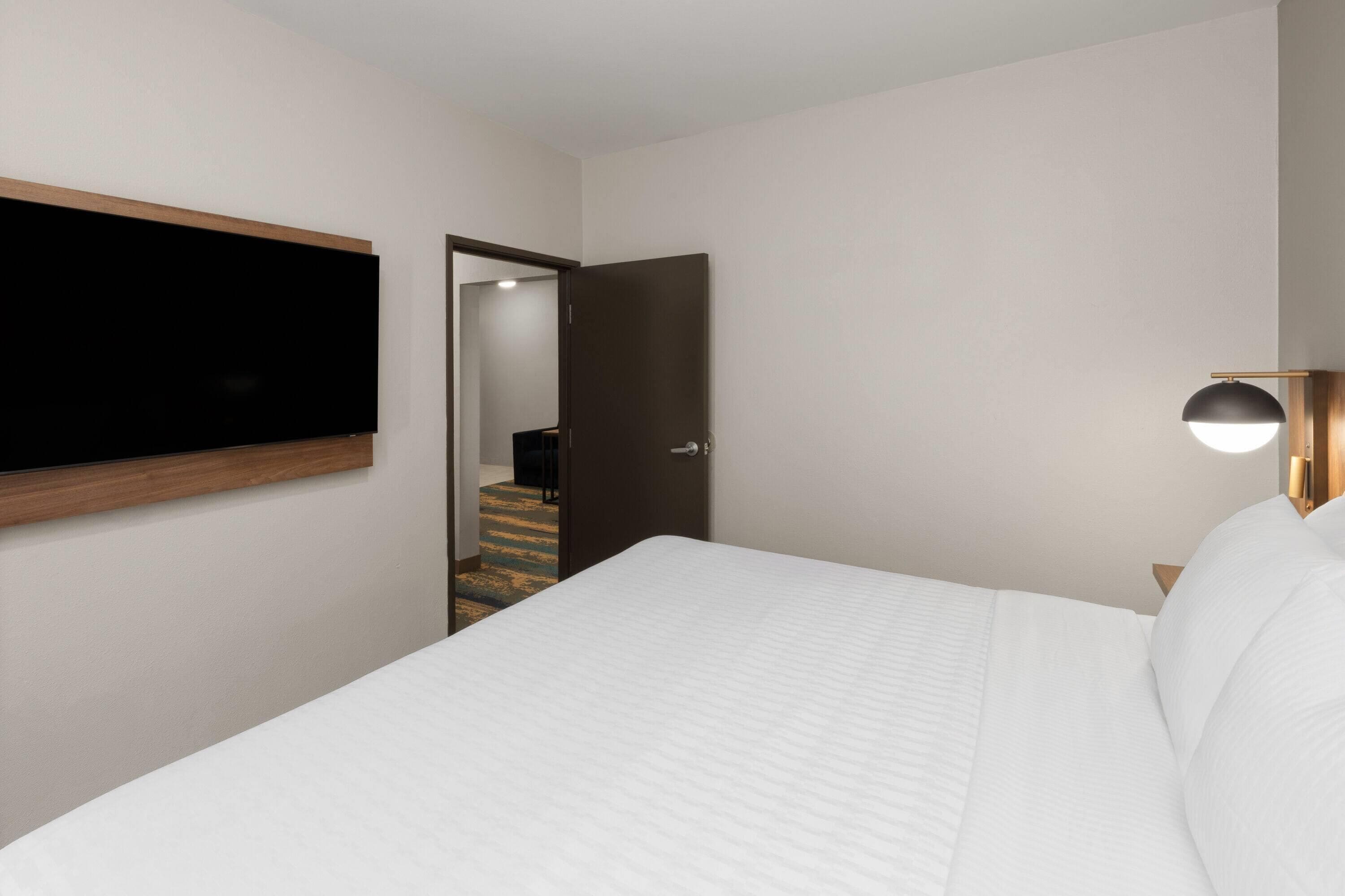 Habitación de hotel con cama, televisor de pantalla plana montado en la pared, puerta que da a un pasillo y lámpara de noche.