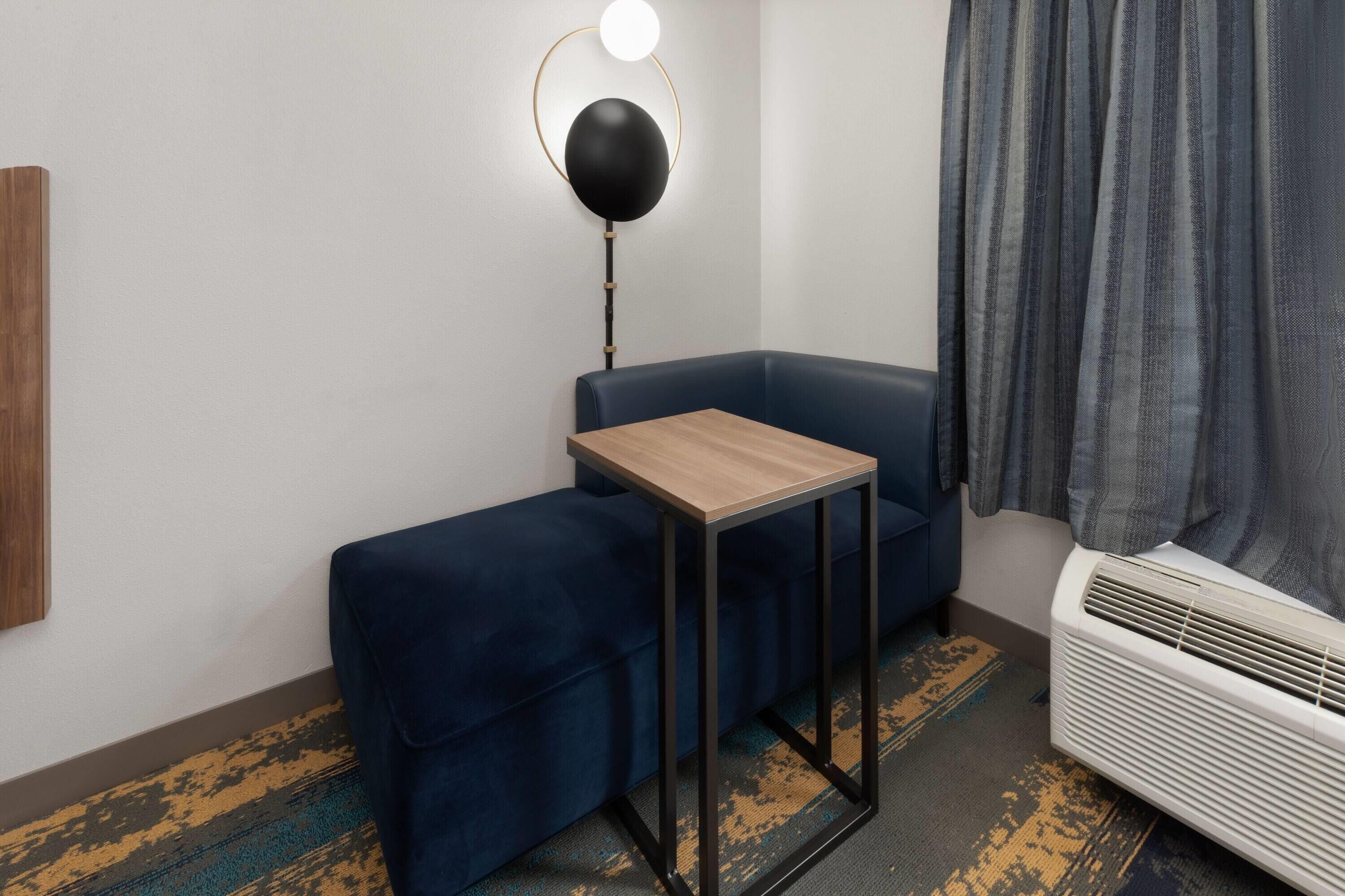 Et hjørneværelse med en blå sofa, et lille træbord, en sort gulvlampe og en hvid aircondition.