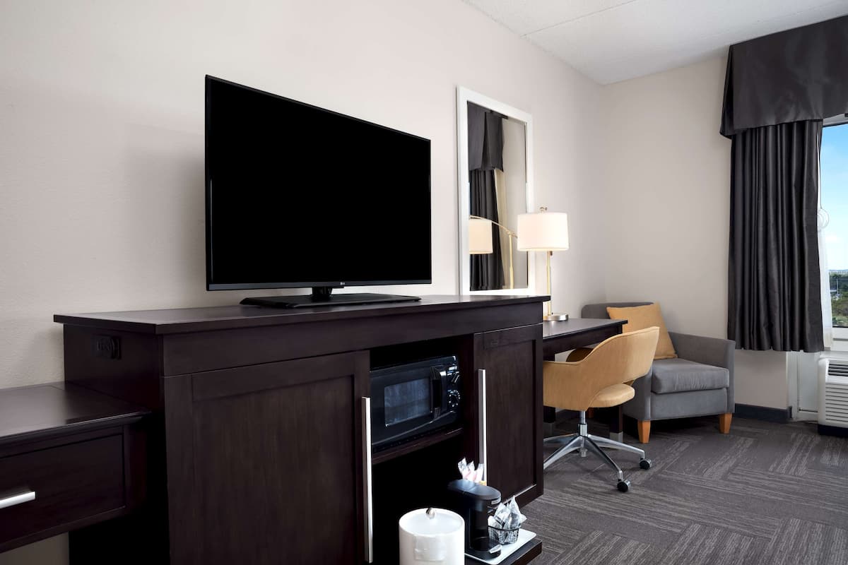Habitación de hotel con un escritorio de madera oscura, televisión de pantalla plana, microondas, una silla, una lámpara y un ventanal con cortinas.